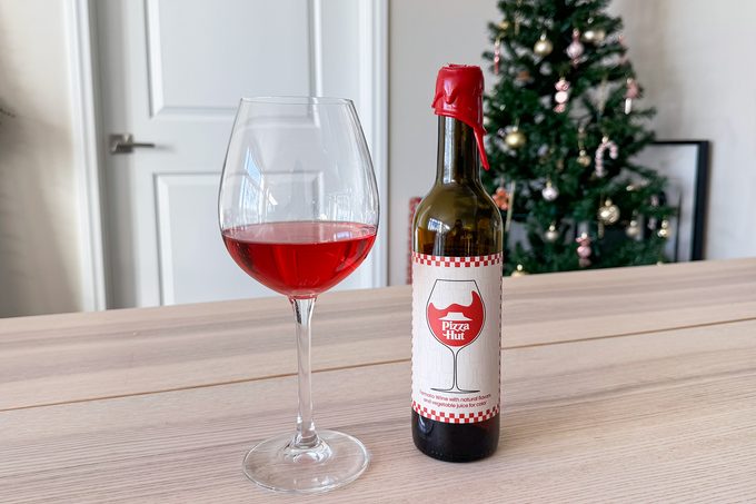 A glass of red wine is next to a bottle with a label featuring a wine glass icon and red accents. A decorated Christmas tree stands in the background. The setting is a light-colored table in front of a closed white door.