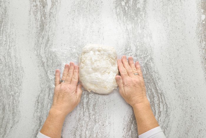 overhead shot of a person's hands wrapping a ball of dough in plastic wrap, the dough is a light color; the hands are on a white textured surface