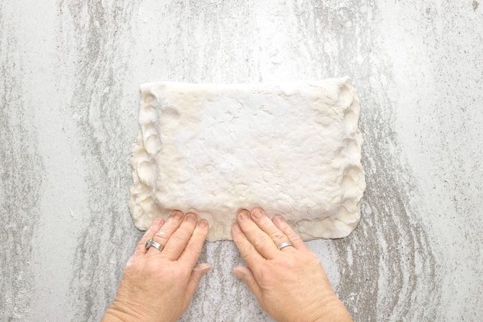 overheda shot of a person's hands, pressing down on a square of dough, possibly pie crust, with both hands; the dough is dusted with flour, it is sitting on a light gray counter or table, there is flour scattered around the dough;
