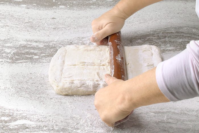 3/4th shot of a person is rolling out dough on a floured surface, using a wooden rolling pin; the dough is a rectangular shape