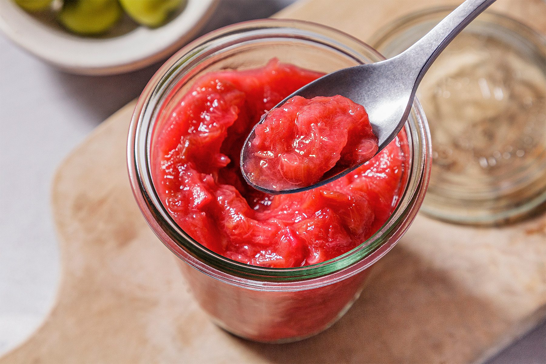 Overhead shot of Rhubarb Compote; in a glass jar; spoon; wooden tray; grey surface;