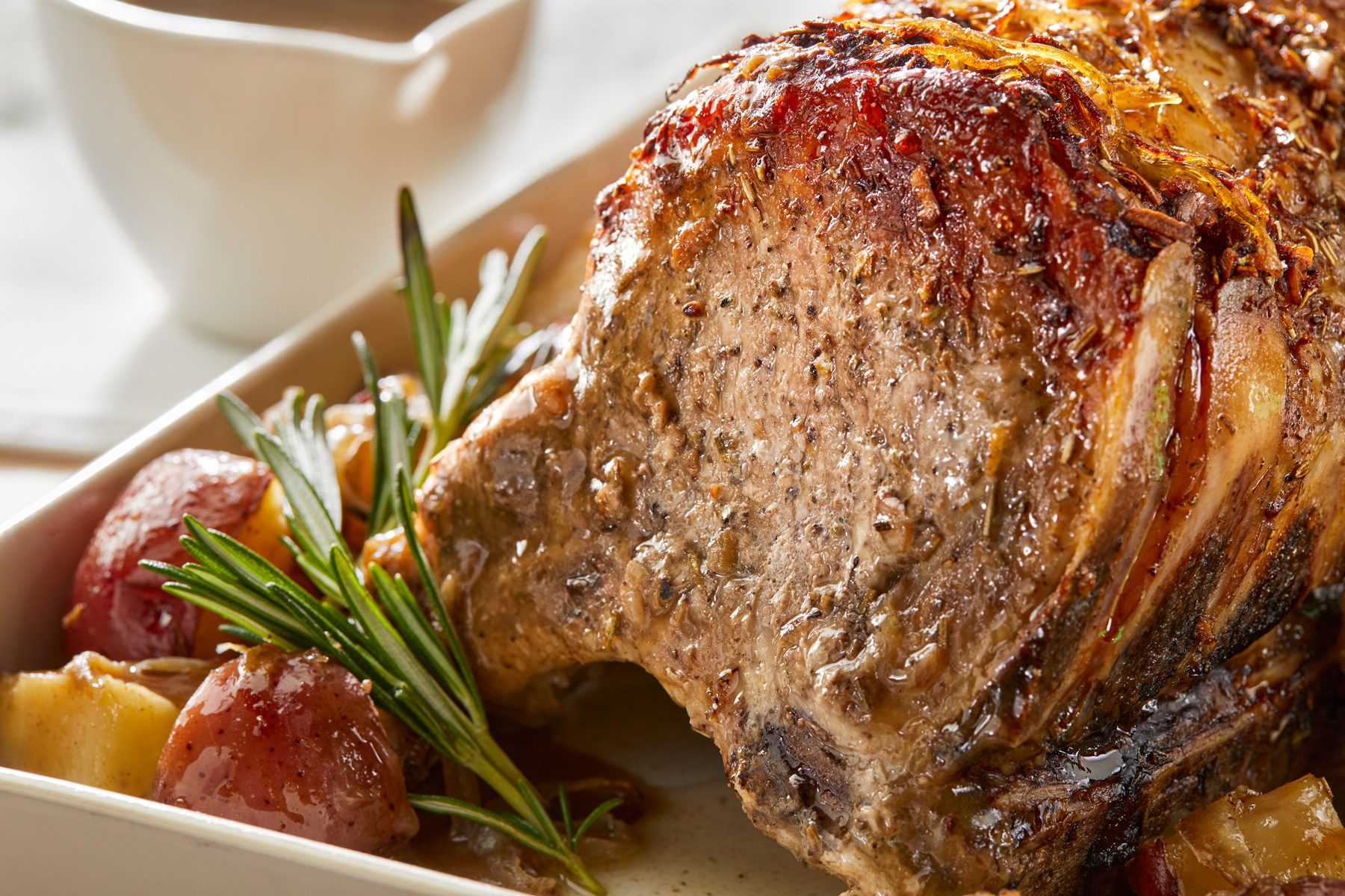 Horizontal Push; close up shot of a roasted pork with potatoes and onions in a white baking dish; there is a sprig of rosemary on top of the pork; there is also a white bowl of gravy next to the baking dish;