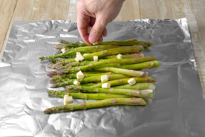 A hand is sprinkling small cubes of butter over a bunch of fresh green asparagus spears arranged on a sheet of aluminum foil. The foil is placed on a wooden surface.