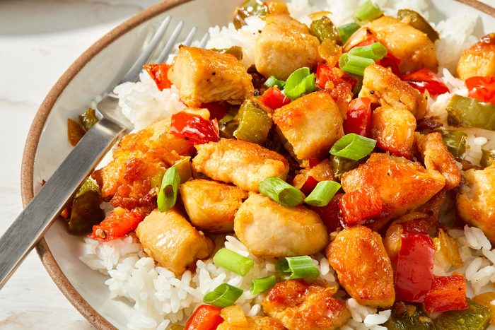 A plate of food that contains Szechuan chicken, rice, green peppers, red peppers and green onions, all of which are cooked in a sauce served with a fork