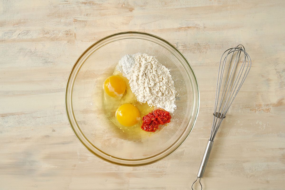 Overhead view of eggs, flour, baking powder, cornstarch, seasoned salt, and harissa in a large bowl for the Taste of Home Sweet Potato Fritters recipe.