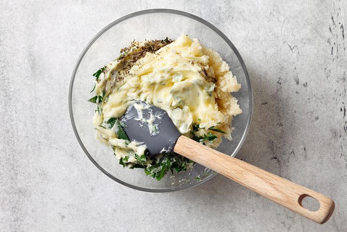 A glass bowl on a gray surface contains mashed potatoes mixed with herbs and spinach. A wooden-handled spatula is stirring the mixture, showcasing a combination of creamy texture and green flecks.