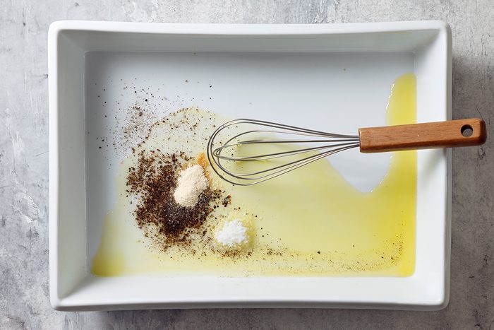 A white rectangular dish contains oil, with a whisk laying in it. Several seasonings are sprinkled next to the whisk, including a light powder and black specks, set against a neutral background.
