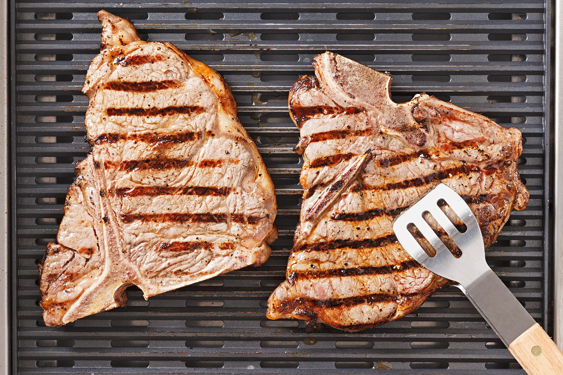 Two T-bone steaks with grill marks are being cooked on a grill. A silver spatula with a wooden handle is resting on the grill near one of the steaks. The surface is dark, contrasting with the brown, juicy meat.