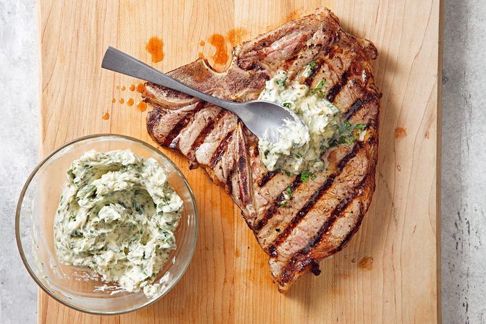 Grilled T-bone steak topped with a dollop of herb butter on a wooden cutting board, accompanied by a bowl filled with more herb butter on the side.