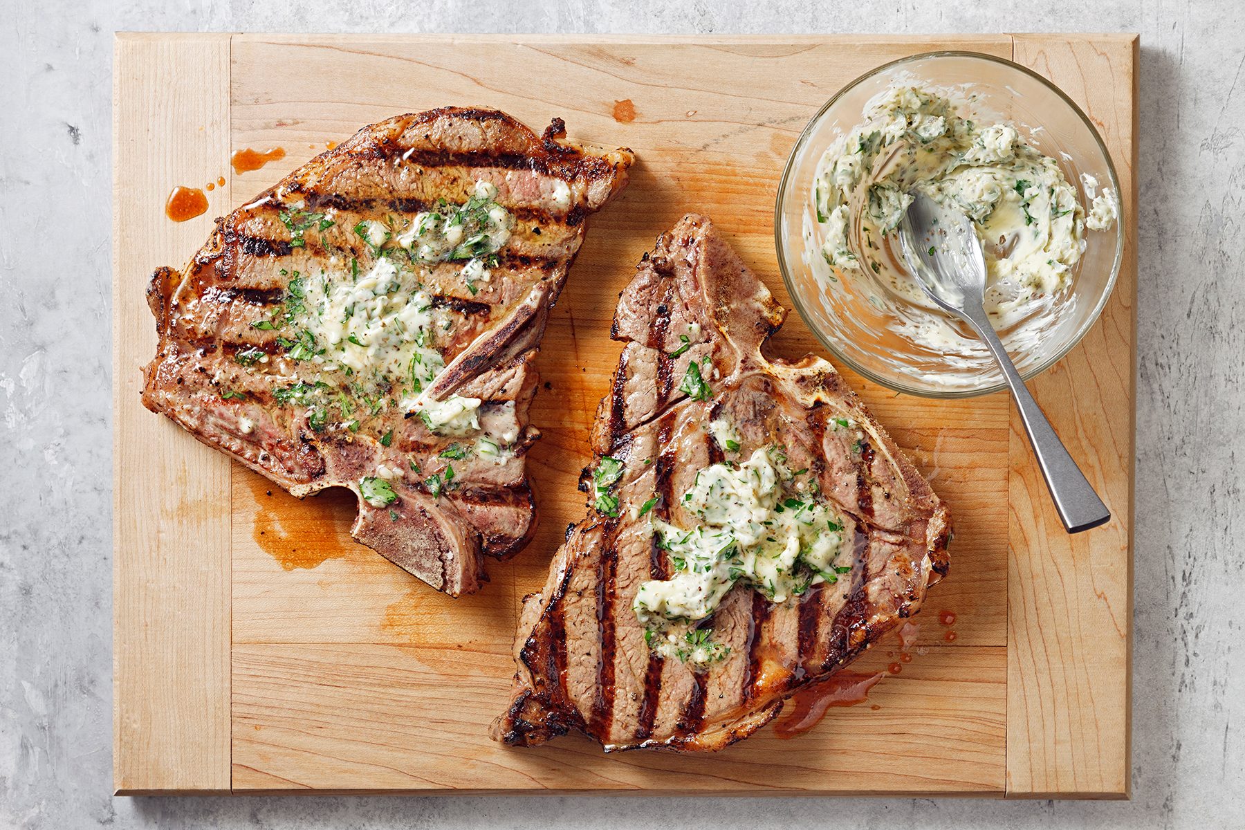 Two grilled T-bone steaks topped with herb butter are on a wooden cutting board. A glass bowl containing more herb butter and a spoon sits beside the steaks. The board rests on a light gray surface.