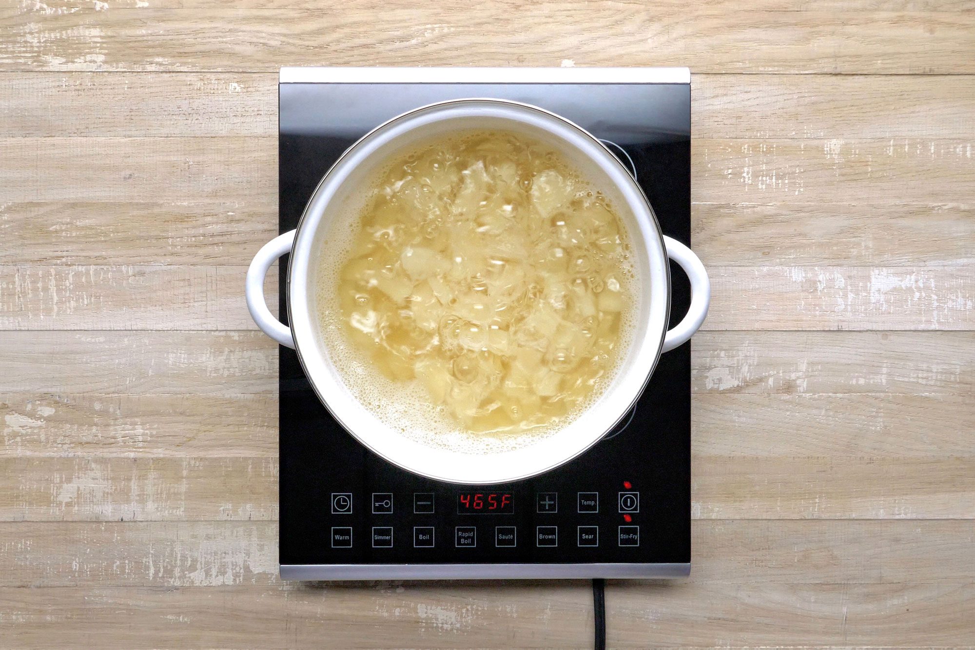 A white pot filled with cooking liquid sits on an electric stovetop. The stovetop has digital controls and is placed on a wooden surface.