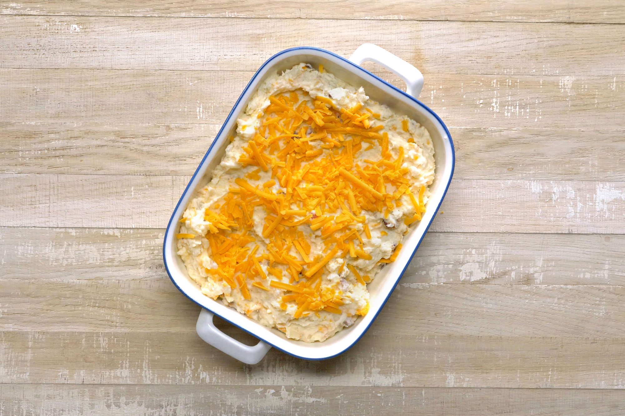 A baked dish in a white rectangular baking pan filled with a cheesy, layered casserole sits on a wooden surface. The top layer is sprinkled with melted orange cheese.
