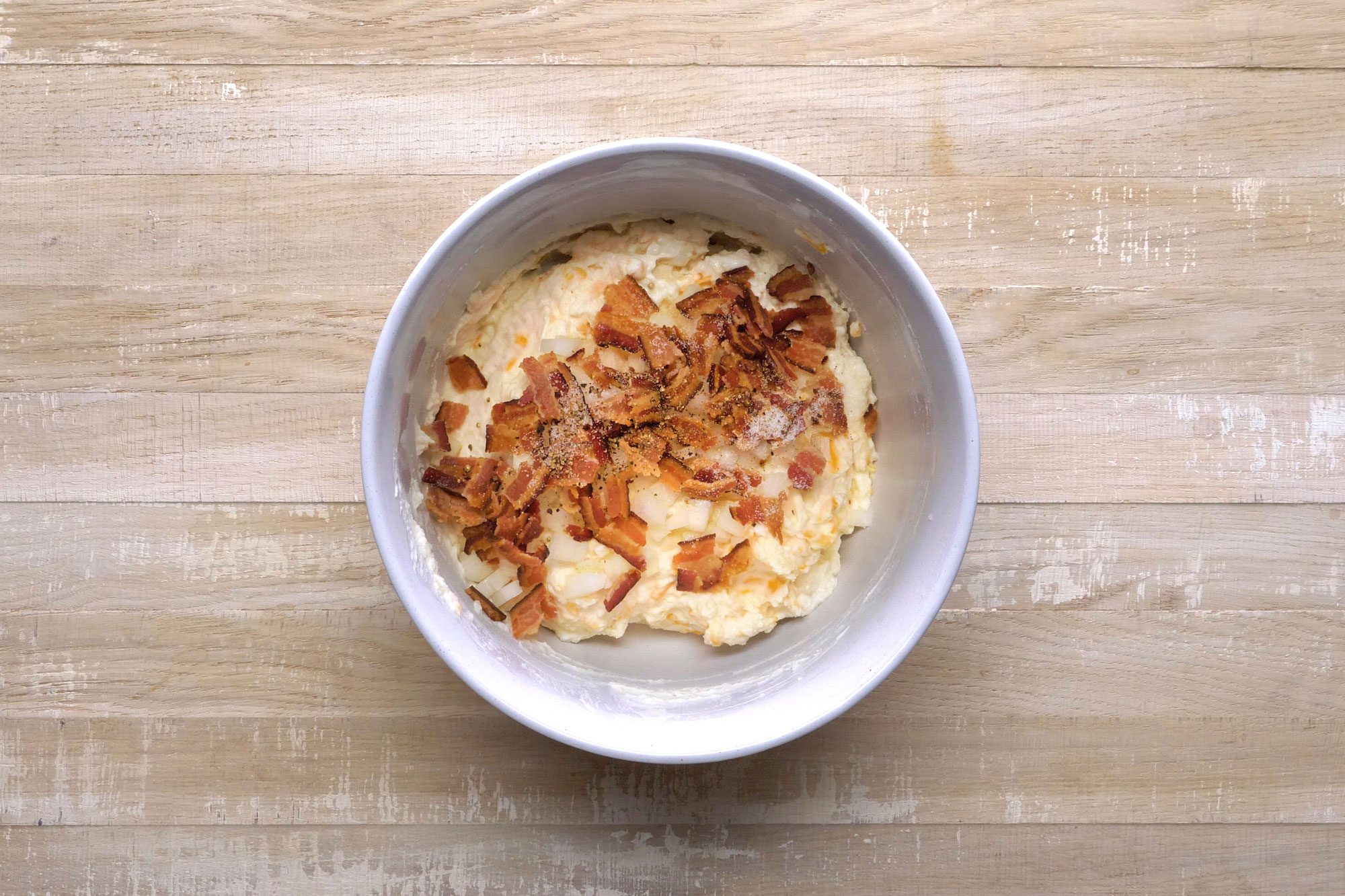 A bowl of creamy mashed potatoes topped with crispy bacon bits on a wooden surface.