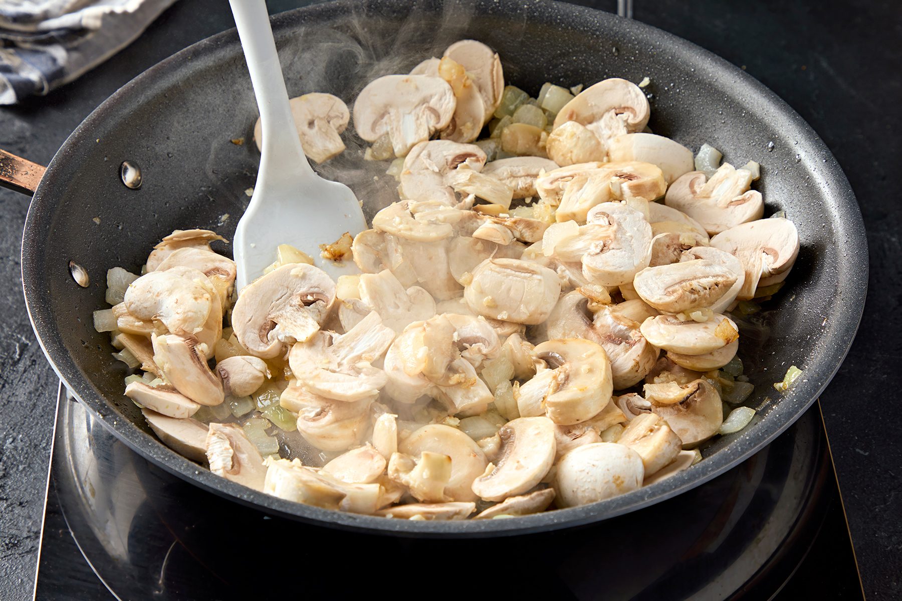 Sliced mushrooms and onions being sautéed in a black frying pan, stirred with a white spatula. Steam rises from the cooking ingredients, indicating heat. The background is dark, highlighting the focus on the pan's contents.
