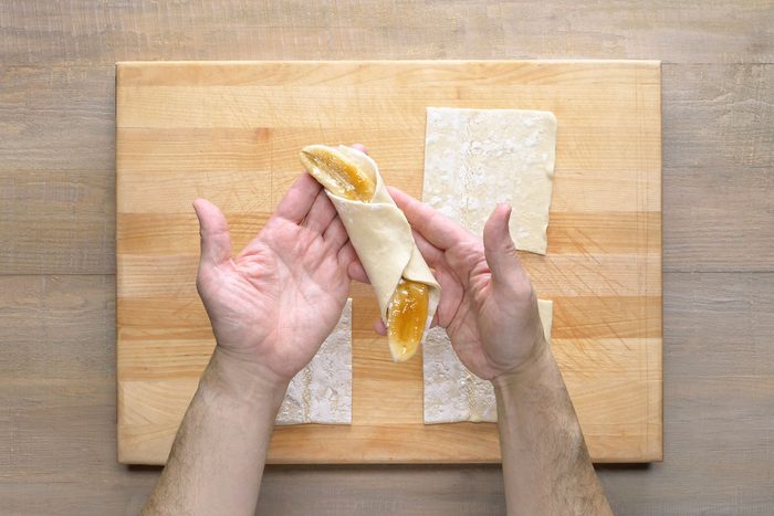 A puff pastry with a filling in it; the pastry is being rolled up with the filling inside; there are several pieces of puff pastry on a wooden cutting board next to the person's hand