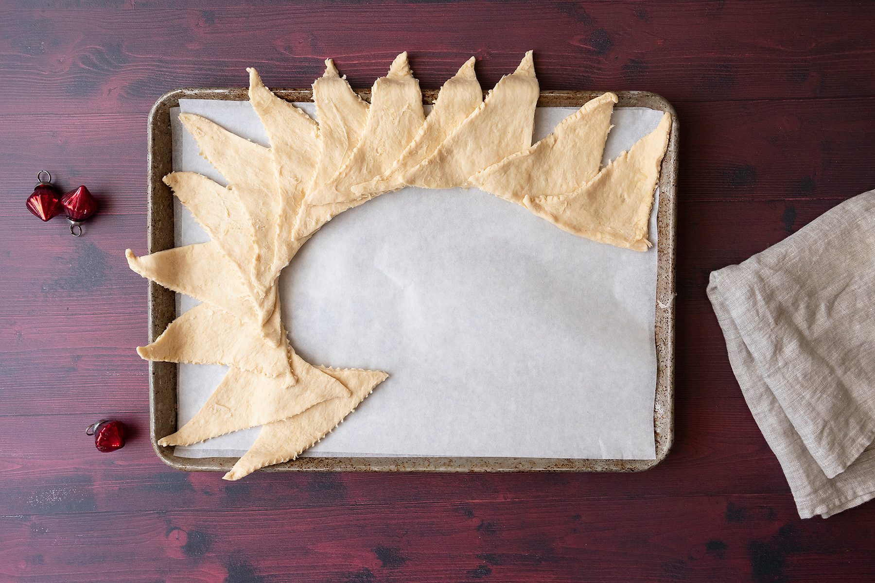 A baking tray lined with parchment paper holds crescent roll dough pieces arranged in a circular pattern resembling a sunburst. To the side, there are two small red ornaments and a folded beige cloth on a dark wooden surface.