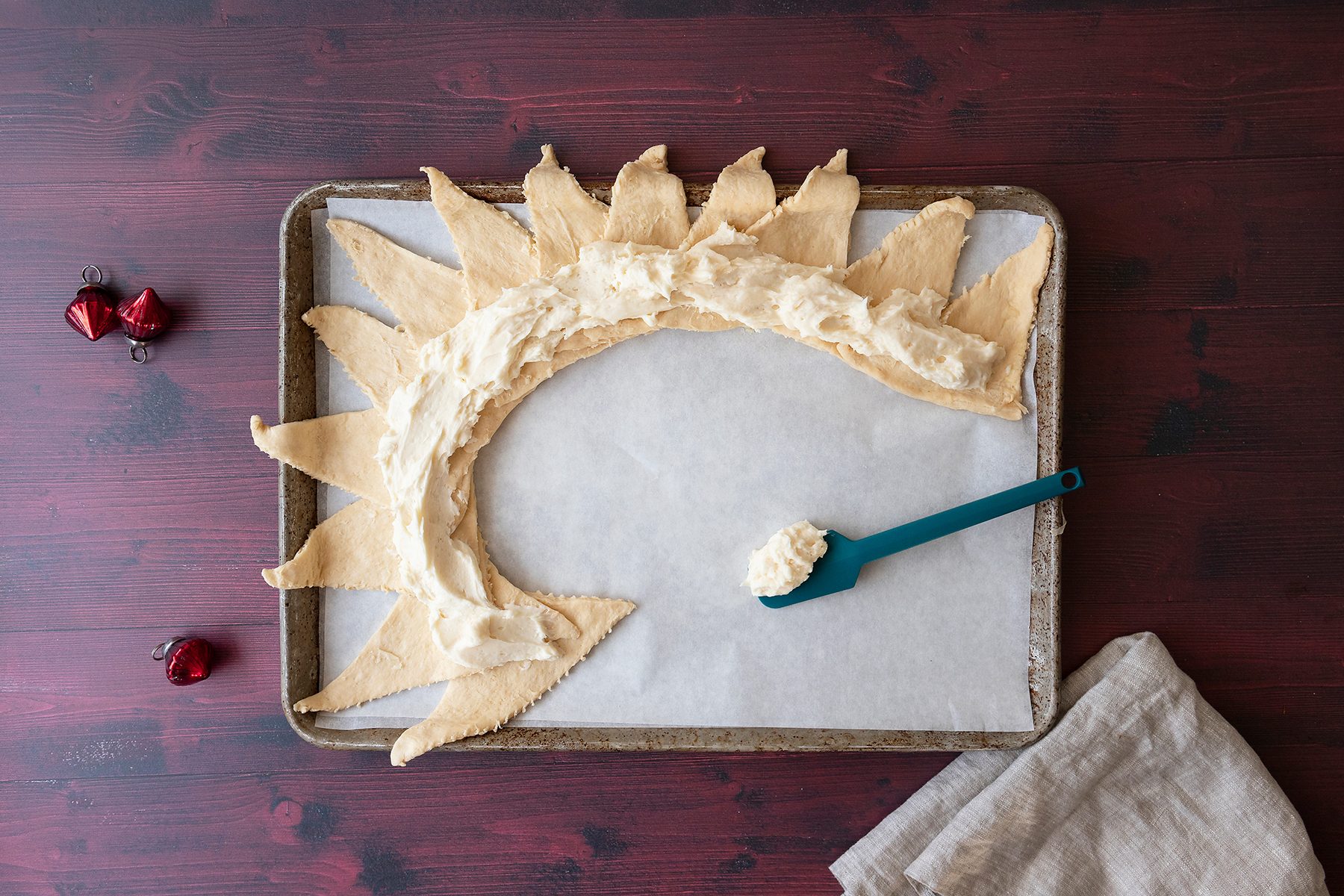 A crescent-shaped pastry with pointed ends is on a baking sheet lined with parchment paper. A blue spoon with cream cheese rests nearby. The background is a dark wooden surface with small red heart decorations. A beige cloth is at the bottom right.