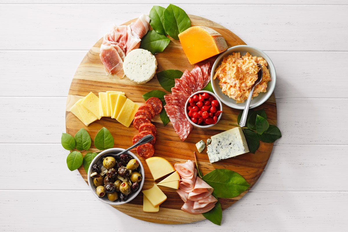A wooden platter with assorted cheeses, cured meats, cherry tomatoes, olives in a bowl, a creamy dip, and leafy greens, arranged aesthetically on a light wooden table.