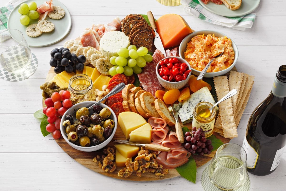 A charcuterie board with assorted cheeses, meats, crackers, grapes, olives, nuts, and small bowls of spreads, arranged on a wooden platter. There are glasses of white wine and small plates in the background on a white table.