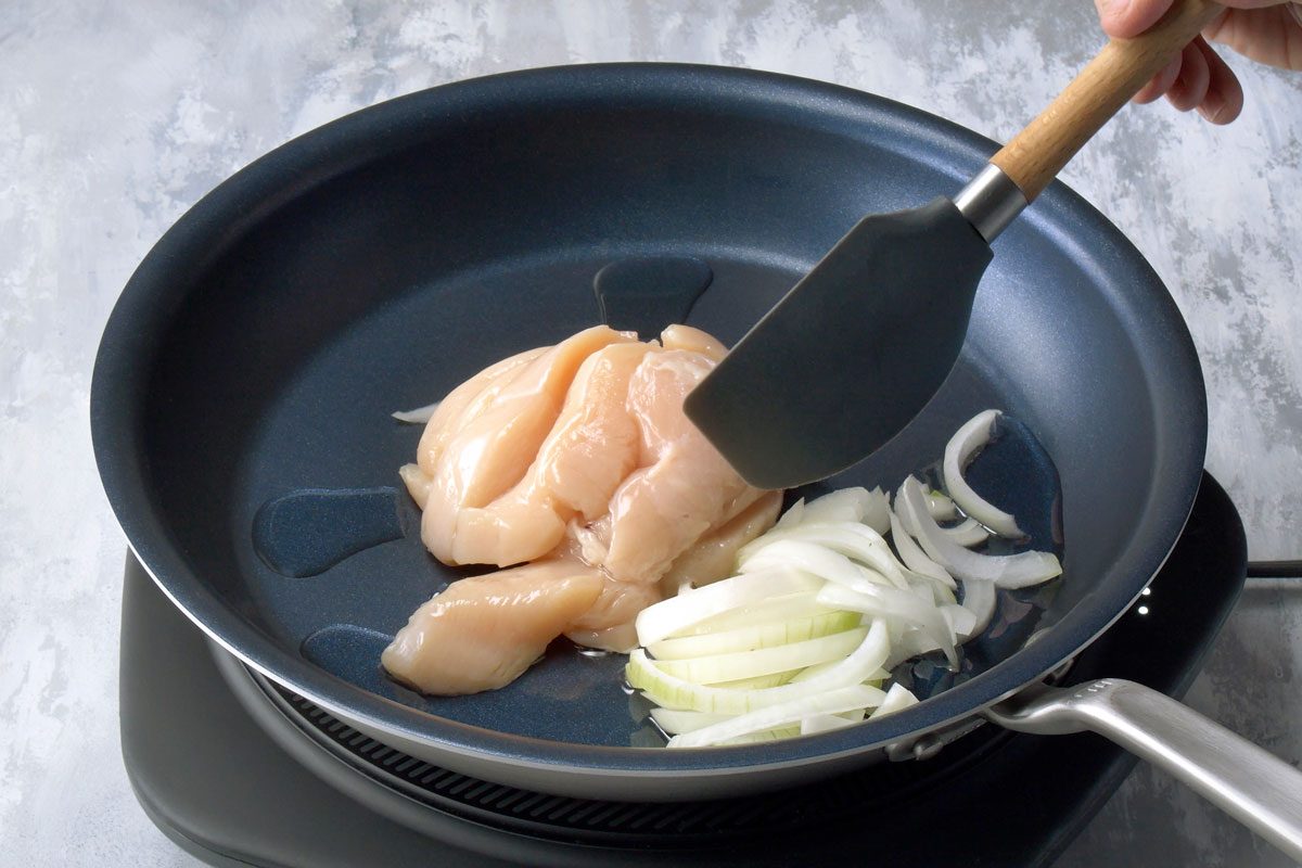 3/4th shot of a black frying pan on a stovetop with raw chicken breast and sliced onions inside, ready for cooking; a hand is holding a spatula with a wooden handle, lifting the chicken to reveal the onions underneath