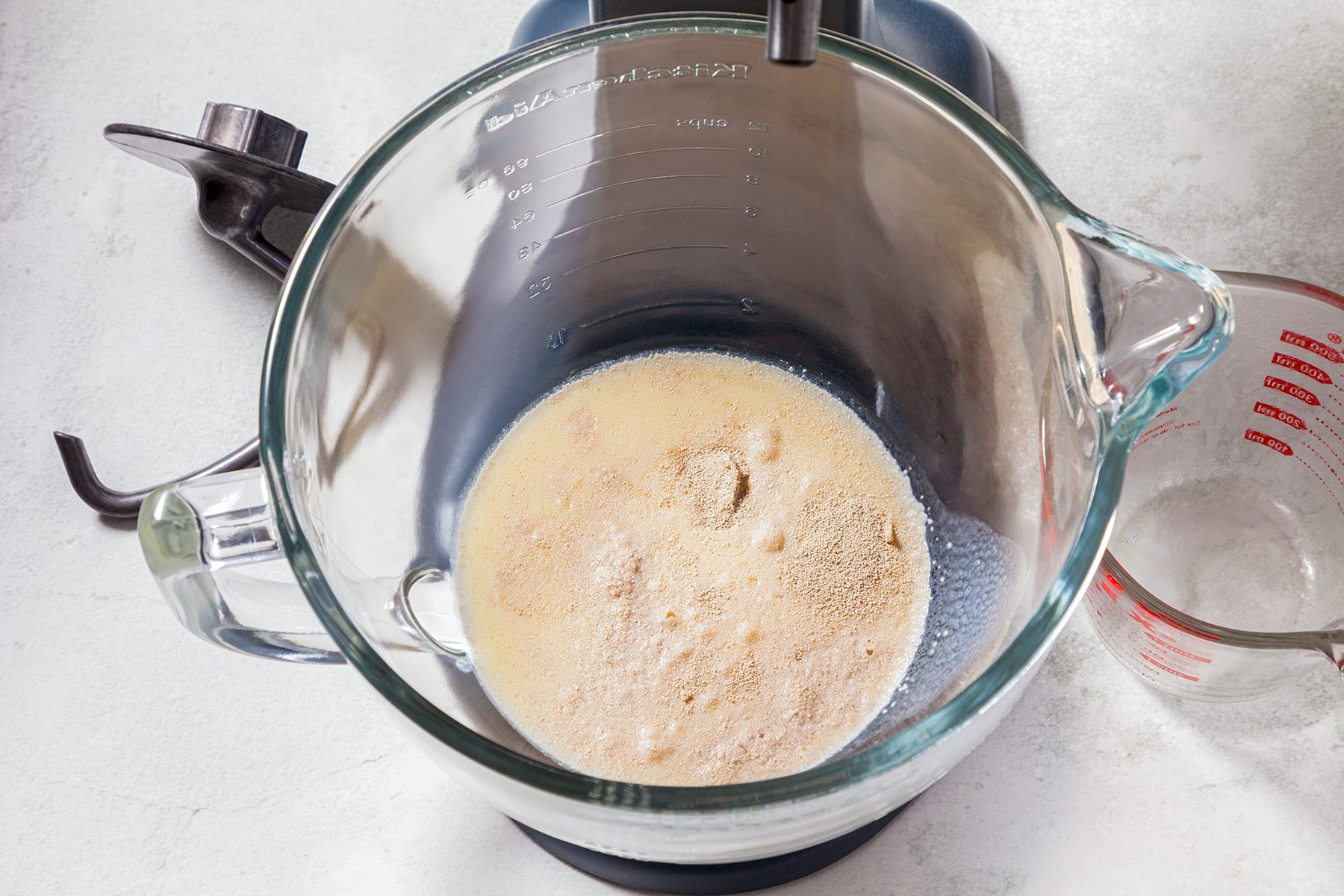 A top view of a blender pitcher containing a beige liquid mixture, resembling batter or dough. It's set on a gray countertop, next to a clear measuring cup with red markings and a black blender base with blades.