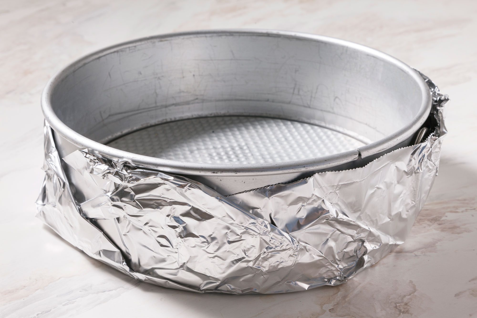 A round metal baking pan wrapped with aluminum foil around the sides, placed on a light marble surface.