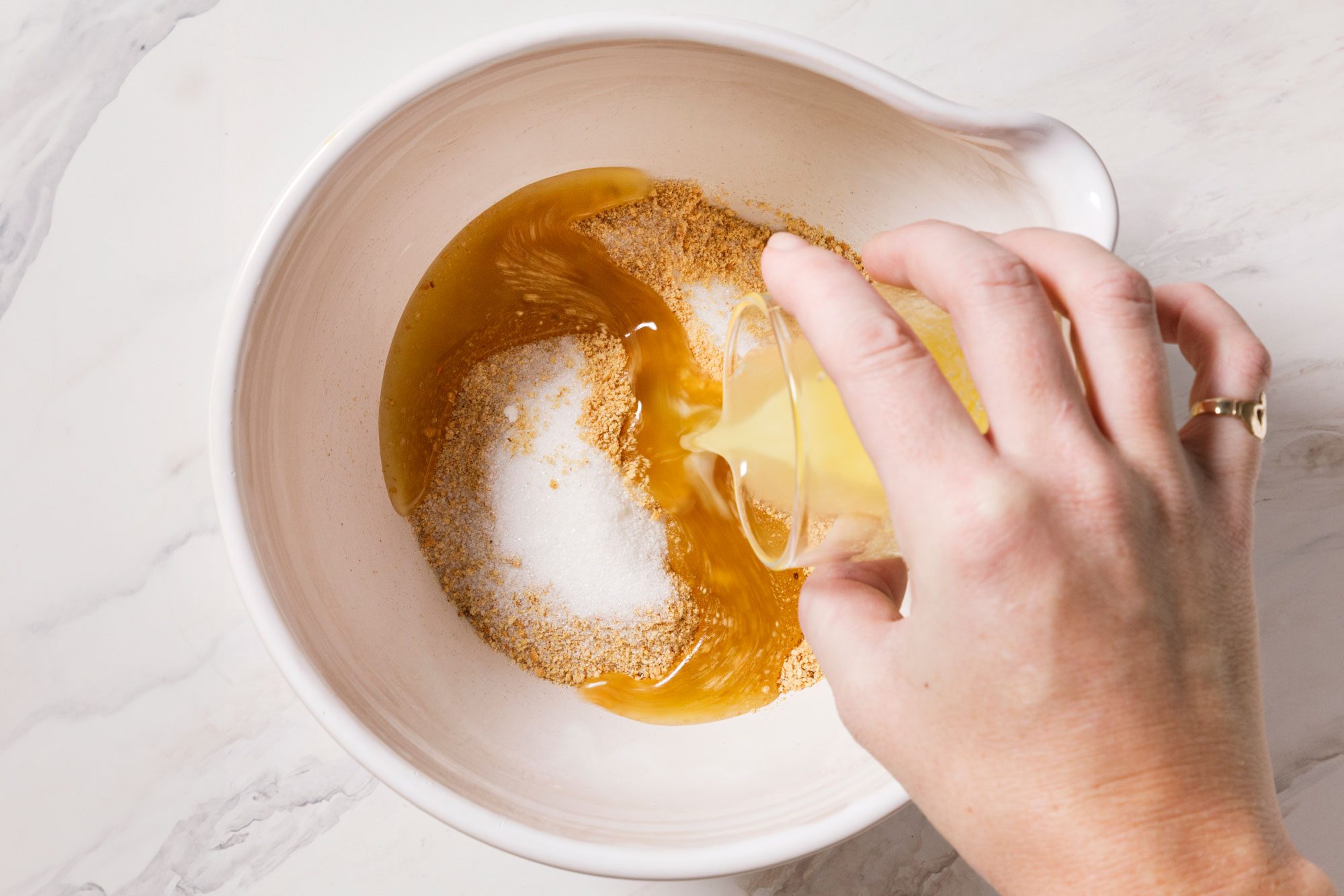 A hand pouring melted butter into a mixing bowl containing sugar and brown sugar, on a marble countertop.