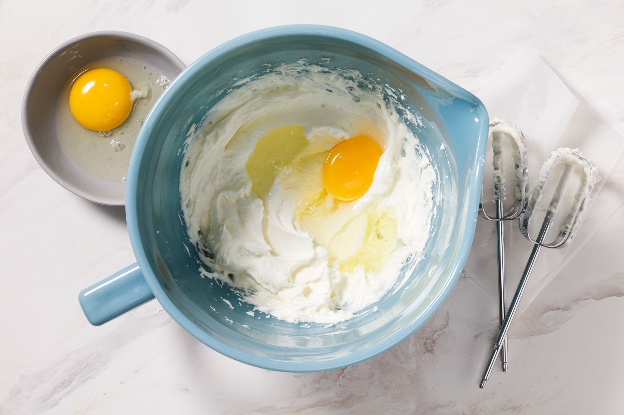 A blue mixing bowl containing cream cheese and an egg on a counter. A separate egg is in a small bowl beside it. Two mixer beaters are placed on the counter nearby.