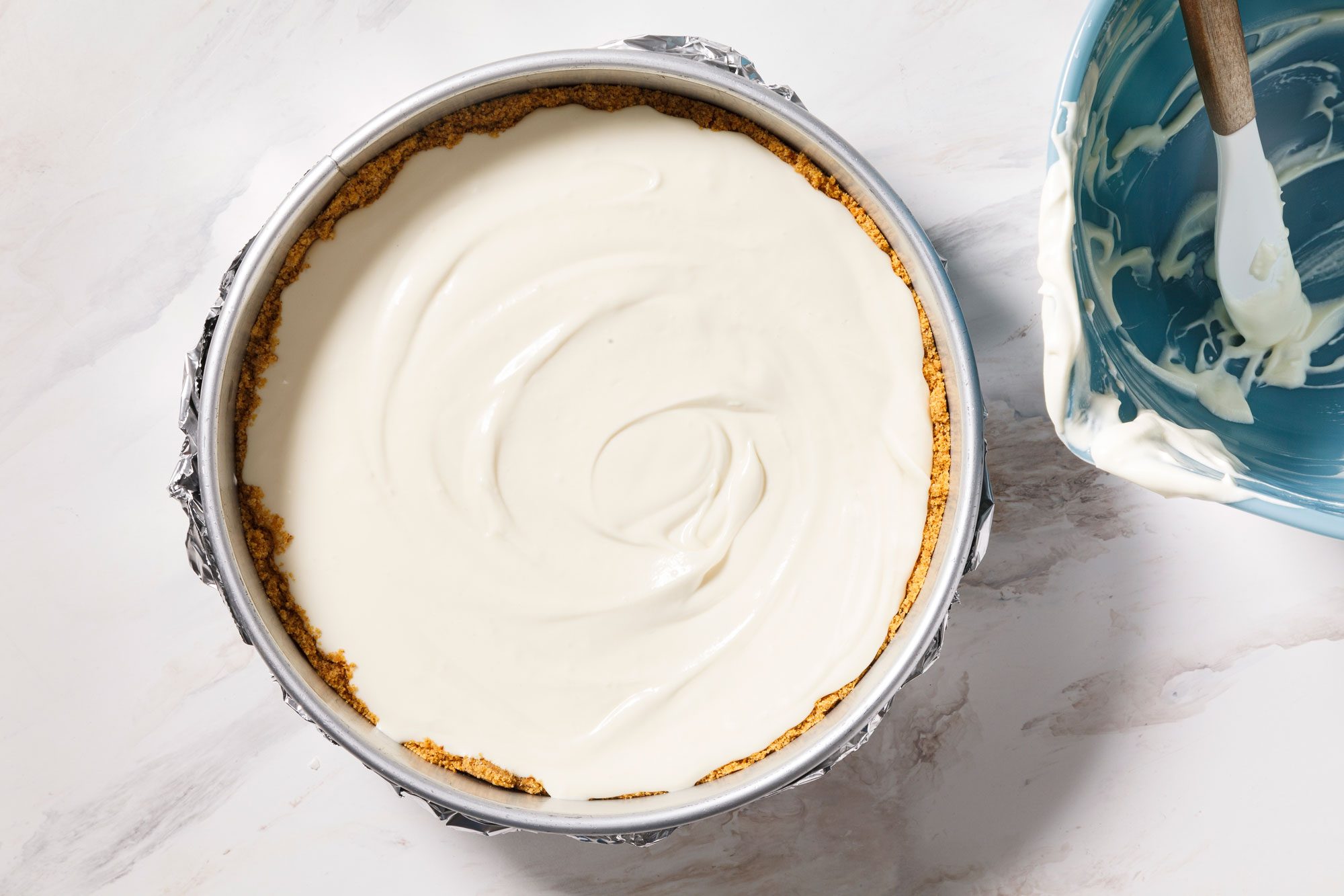 A top view of a cheesecake with a smooth, creamy filling in a round pan. A blue bowl with a spatula and some leftover cream is on the side, all placed on a white marble surface.
