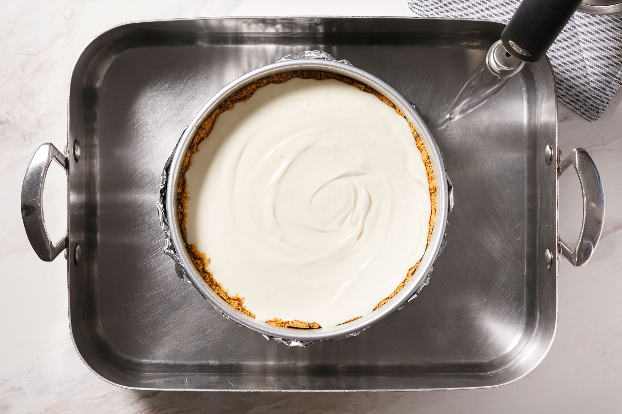 A round cake pan with a cheesecake inside sits in a large, shallow roasting pan filled with water, on a kitchen countertop. A striped towel is partially visible beside the pans.