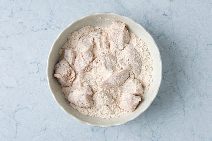 Chicken pieces coated in flour are placed in a white bowl on a light blue marble surface.