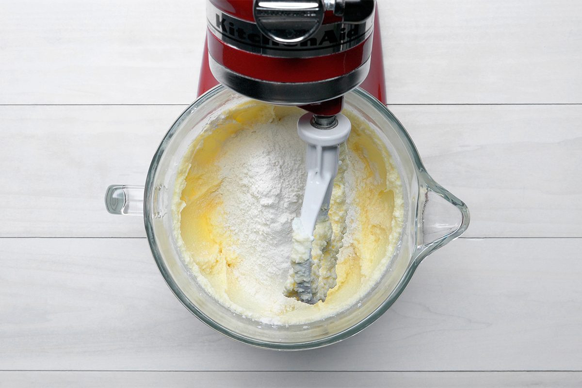 A red stand mixer with a glass bowl is mixing creamy yellow batter and white flour. The bowl sits on a white wooden surface.