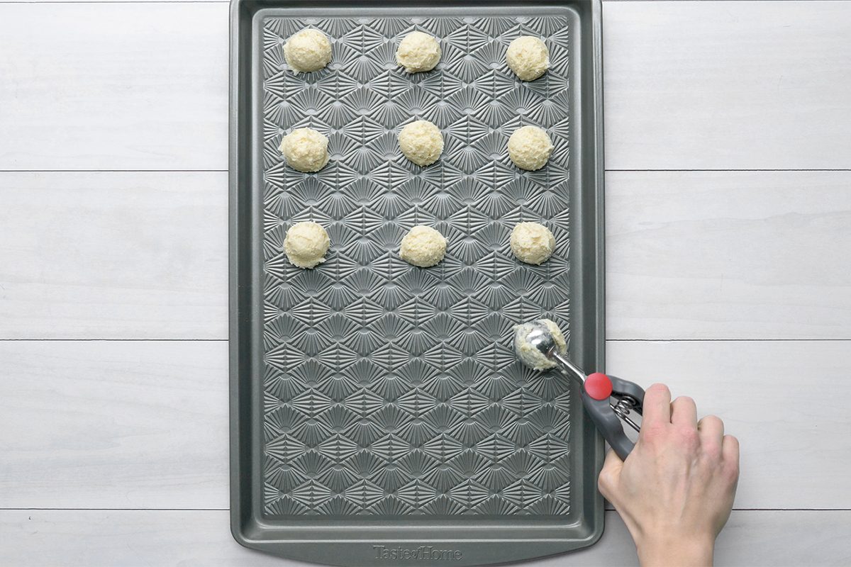 A baking tray with nine evenly spaced cookie dough scoops on a patterned surface. A hand holding a cookie scoop is adding more dough. The tray is on a light-colored wooden table.