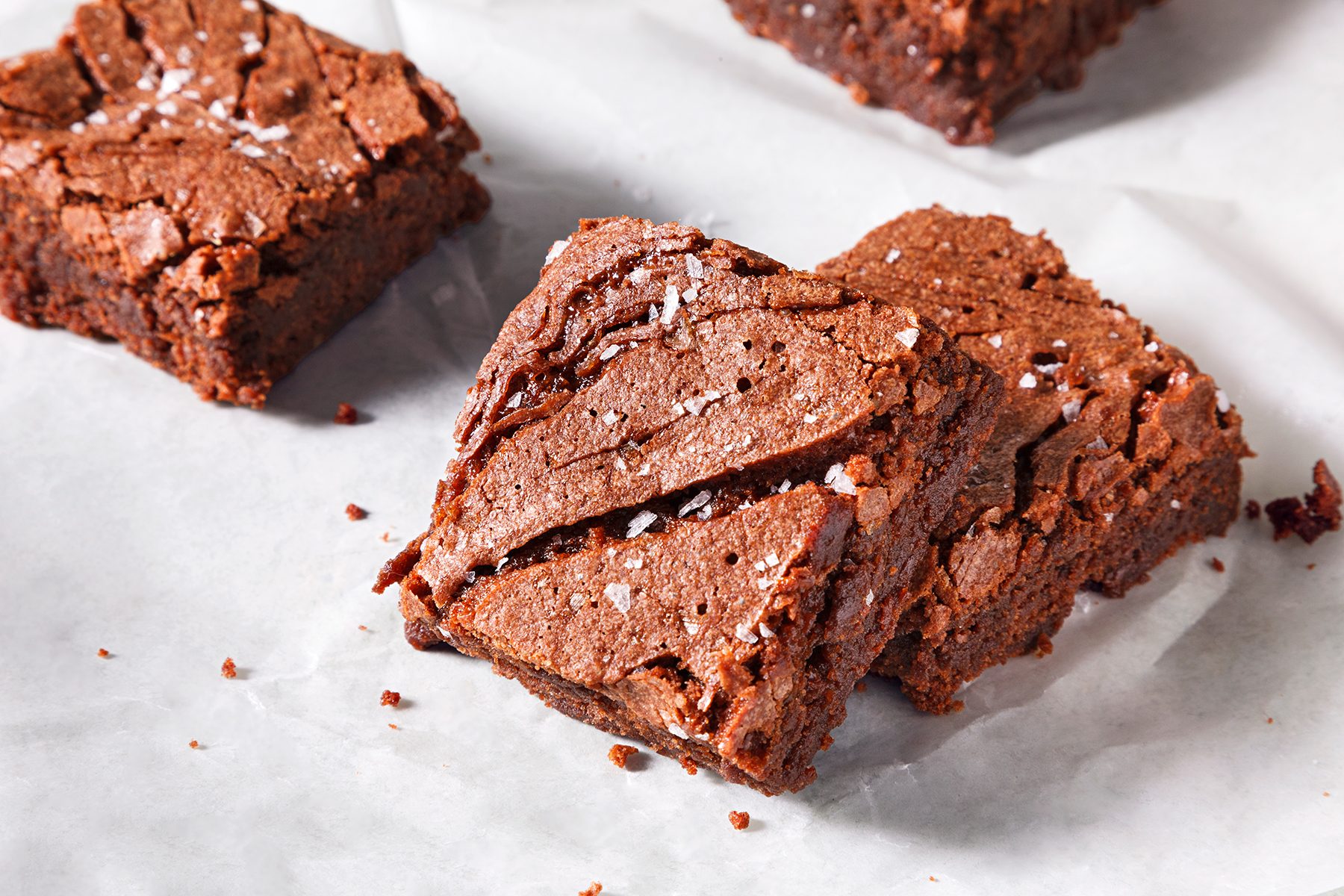 Close-up of two square chocolate brownies on parchment paper. The brownies have a slightly cracked top and are sprinkled with coarse sea salt, highlighting their rich and fudgy texture.