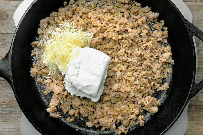 Ground meat in a skillet with a pile of shredded cheese and a scoop of cream cheese on top.