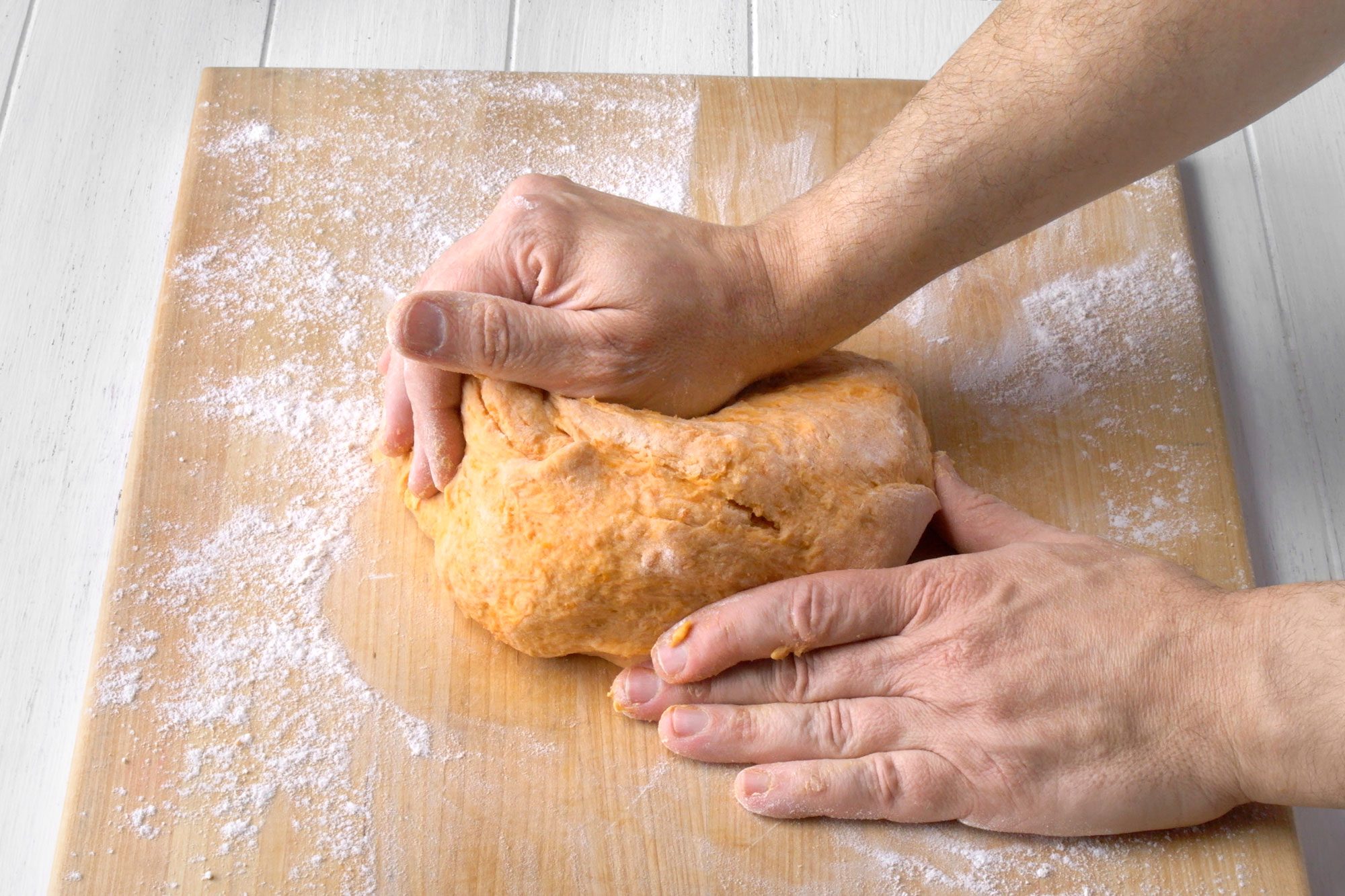 3/4th shot of a person kneading dough on a wooden board; the board is covered in a layer of flour; the dough is orange in color and the person's hands are covered in flour; the person is using both hands to knead the dough, one hand is on top of the dough and the other hand is underneath; the background is a white wooden surface