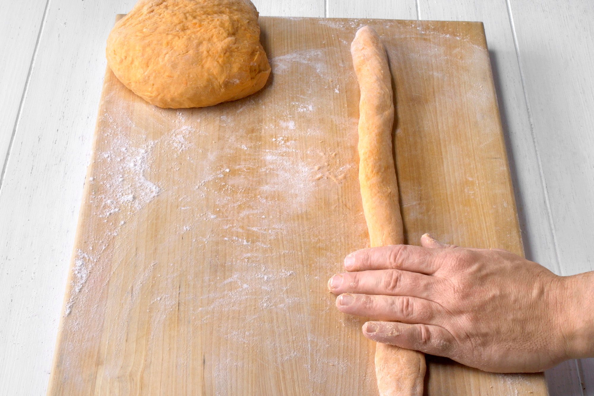 3/4th shot of a hand rolling out a long piece of orange dough on a wooden cutting board, the board is dusted with white flour, there is a round ball of the same orange dough to the left of the hand; the hand is holding the dough in place as it is being rolled out, the dough is smooth and uniform in texture; the background is a white wooden surface with a few light wood grain markings