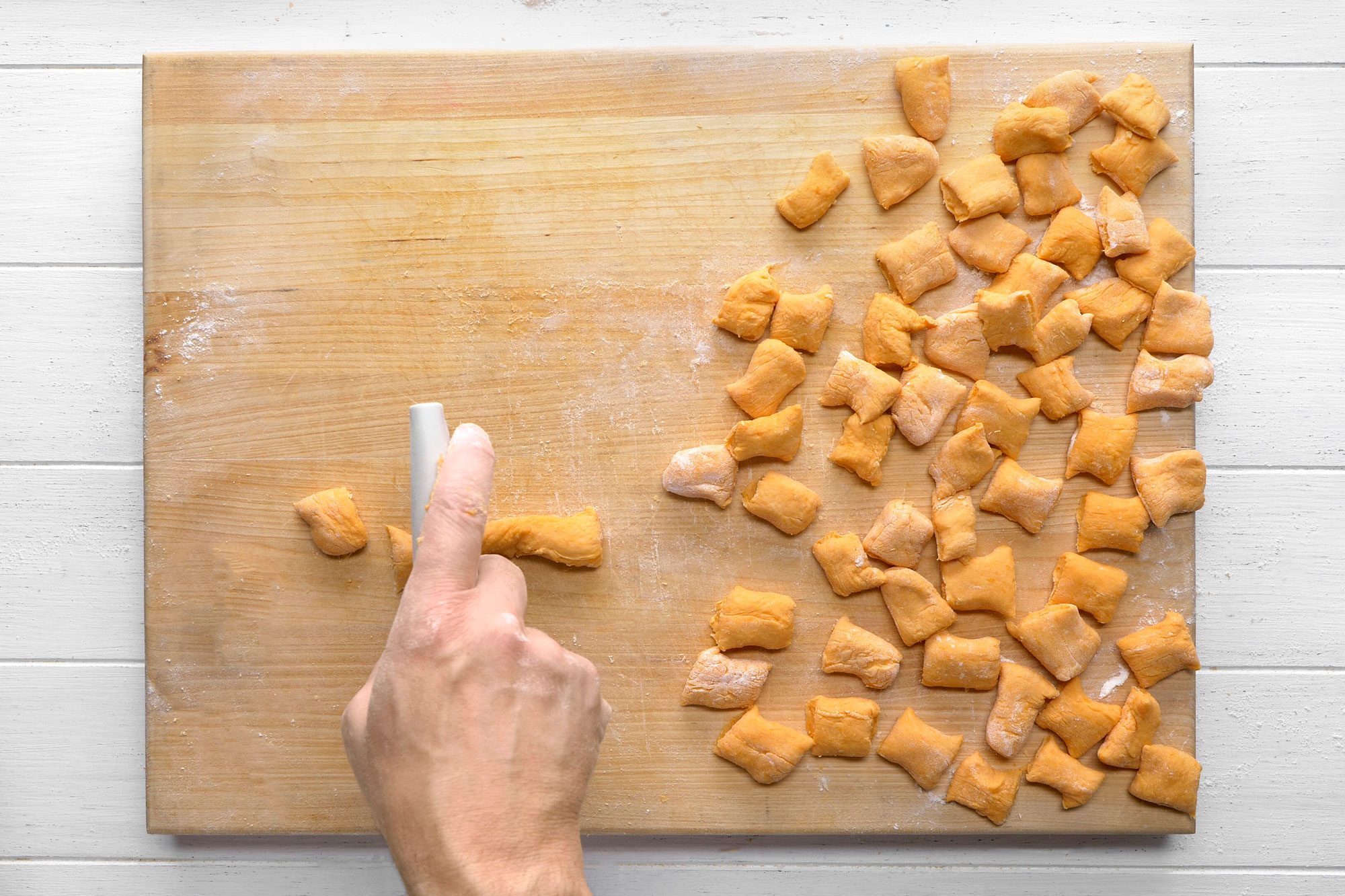 overhead shot of a person making gnocchi on a wooden cutting board; there is a hand pressing down on a long piece of gnocchi dough with a white tool; there are many gnocchi pieces already made, dusted with flour, scattered on the cutting board; the cutting board sits on a white wood background