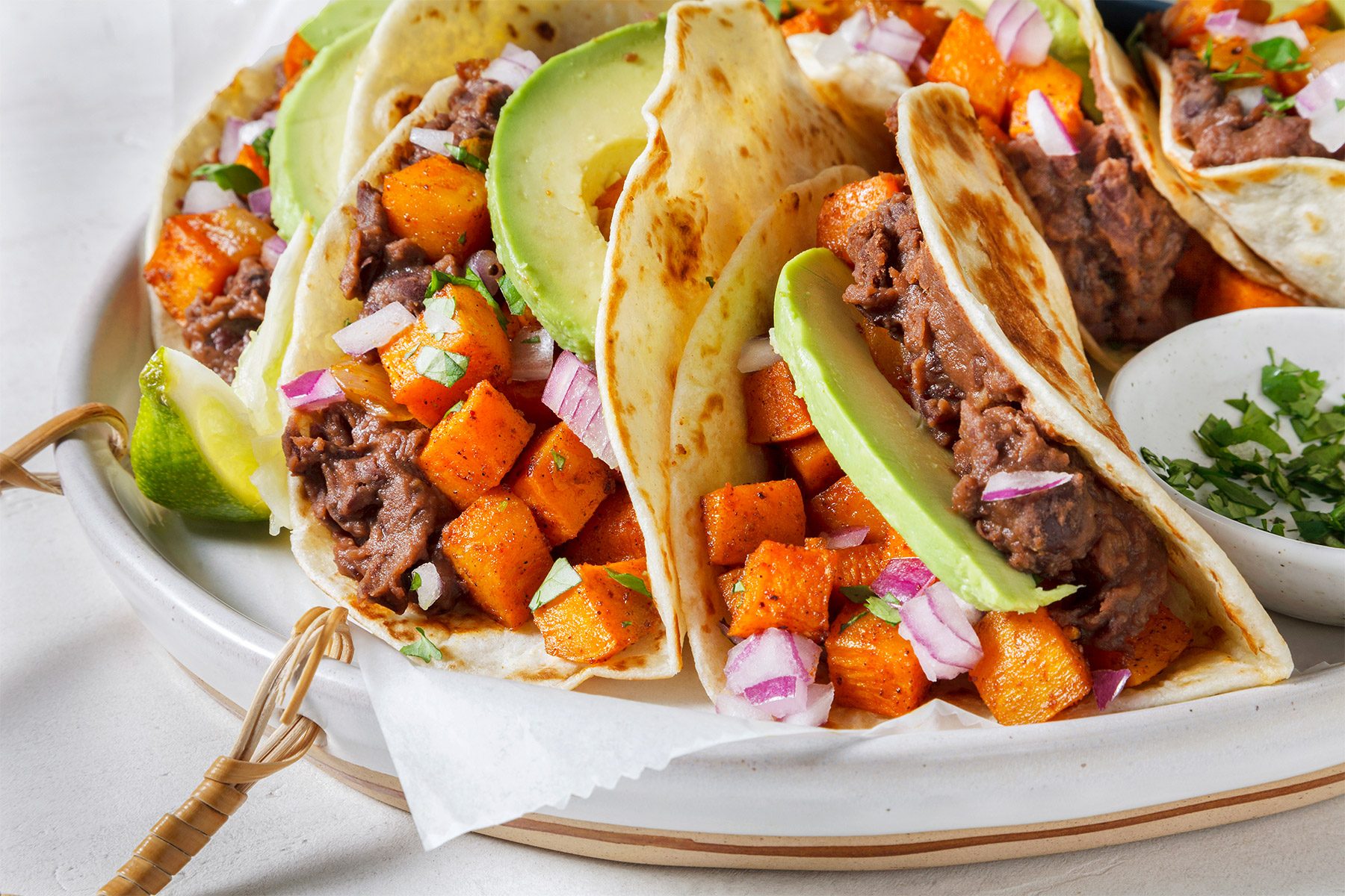 Close-up of three soft tacos on a white plate. Each taco is filled with black beans, sliced avocado, diced red onion, and roasted sweet potatoes. A garnish of fresh cilantro is visible beside the dish.