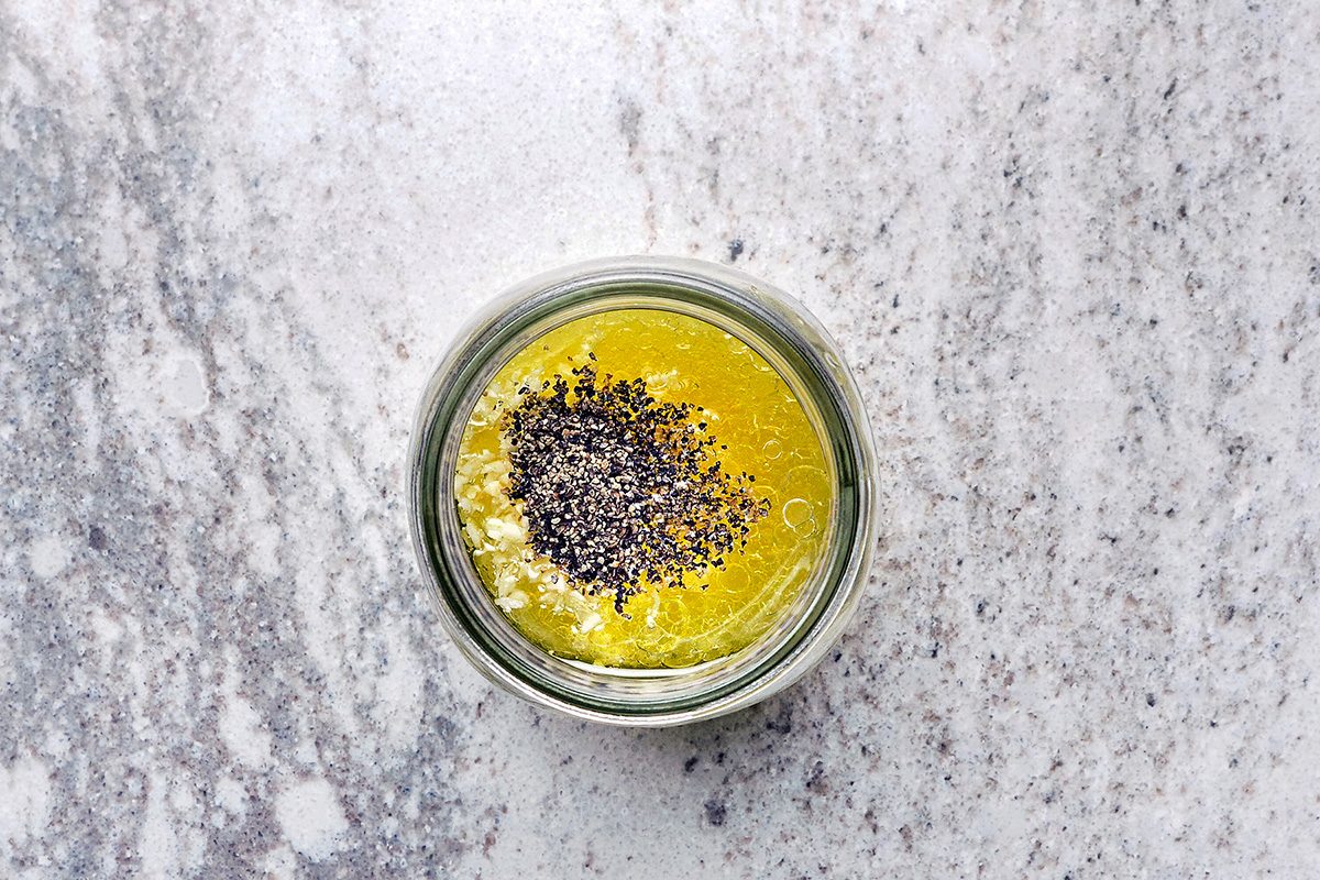 A glass jar filled with yellow oil and sprinkled black seeds on top, set on a textured gray surface.