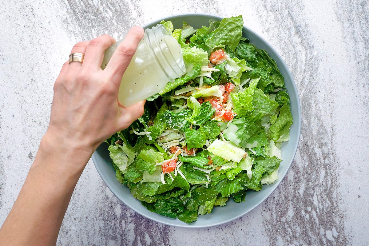 A hand pouring dressing from a jar onto a fresh salad with lettuce, tomatoes, and shredded cheese in a blue bowl. The background has a marble texture.