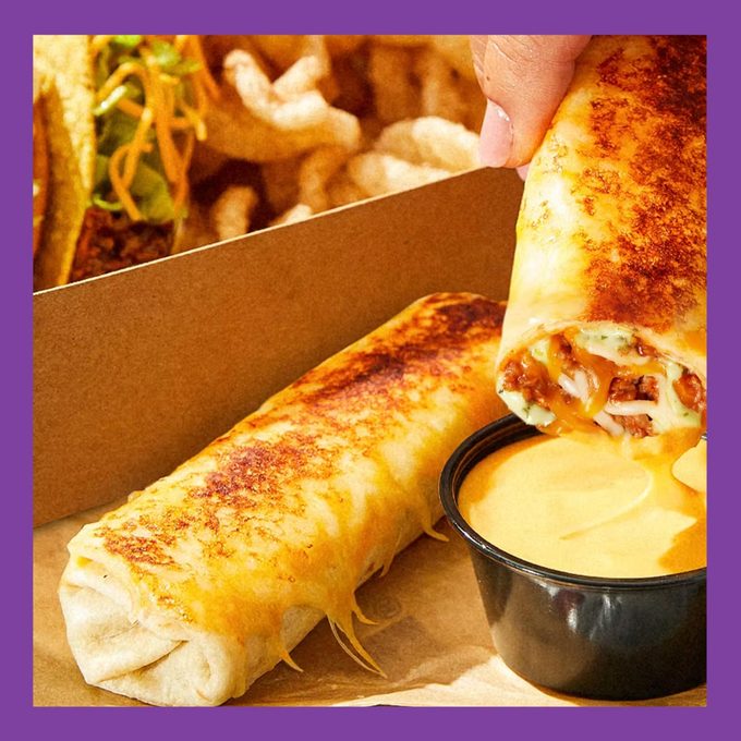 Taco Bell's upcoming Cheesy Dipping Burrito featured with a cup of queso and a box of tacos in the background