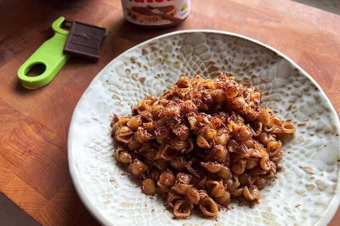 White bowl of chocolate pasta shells on a wooden cutting board with a green peeler, chocolate square, and jar of Nutella in the background