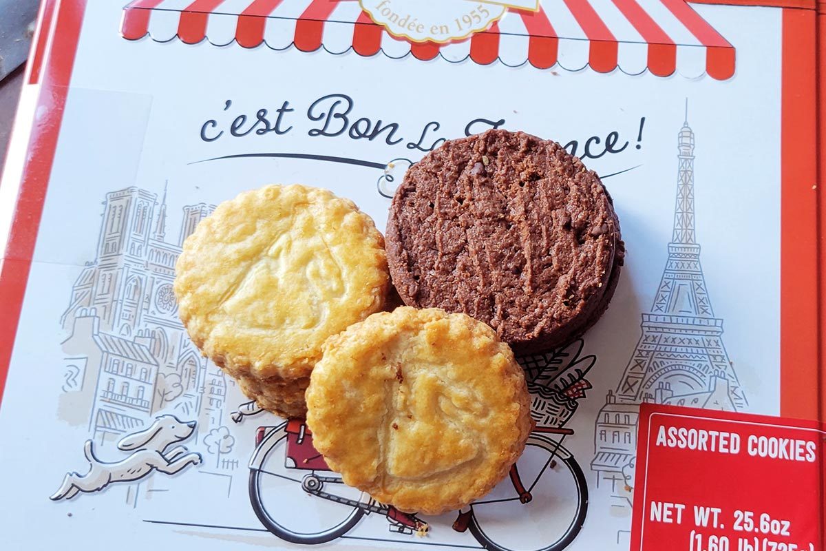 Three round cookies - one light tan, one beige, and one dark brown, sit on top of a french-themed red-and-white box