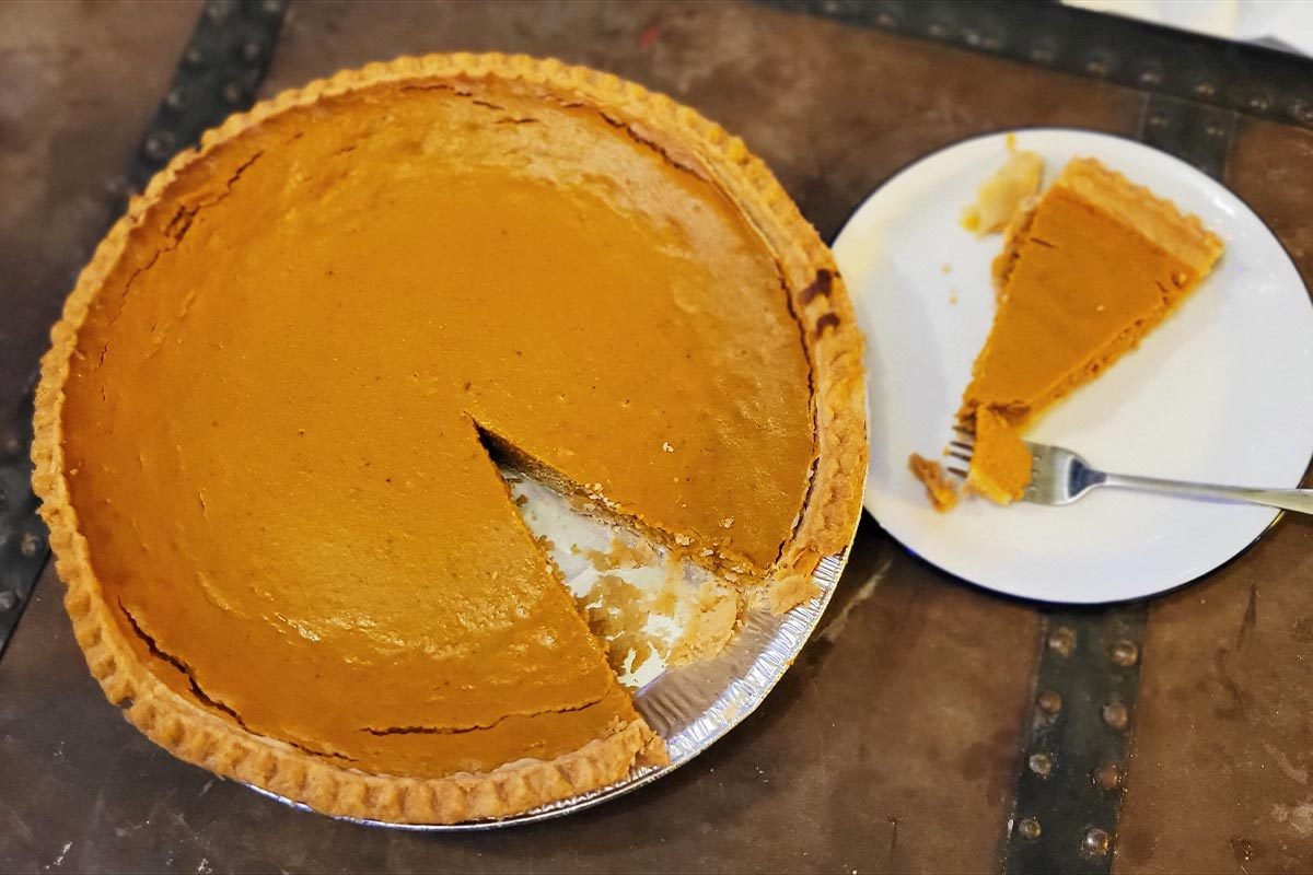 A large pumpkin pie with a slice taken out of it, with the slice on a small white plate with a fork next to it
