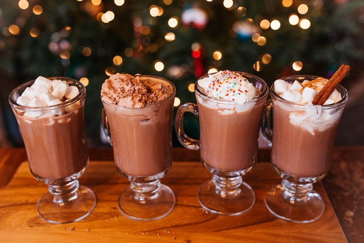 Four glass mugs of hot chocolate with different toppings on a wooden board in front of a festive holiday light background