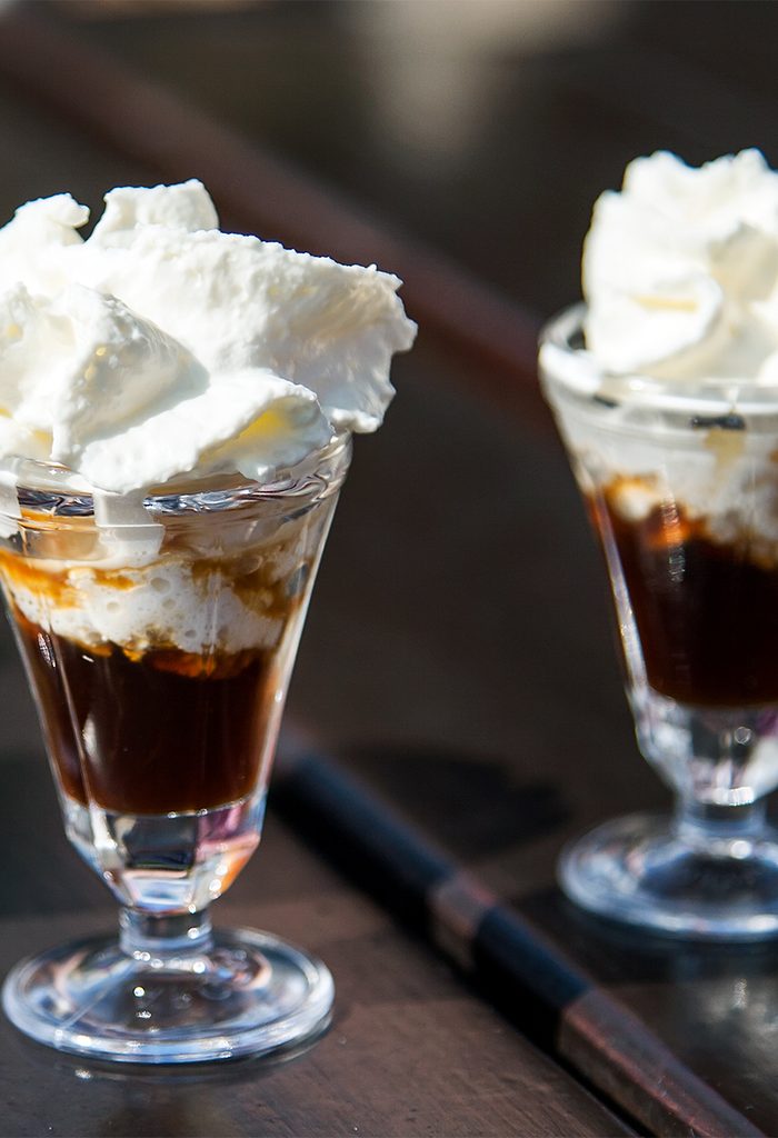 Two glass dessert cups filled with layers of dark espresso and topped with generous swirls of whipped cream, set on a wooden surface.