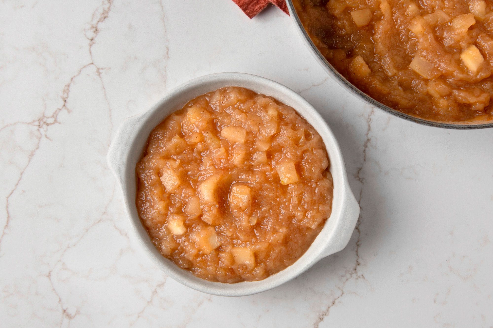 Overhead shot of a bowl of applesauce on a marble countertop, the bowl is white and has a rounded shape