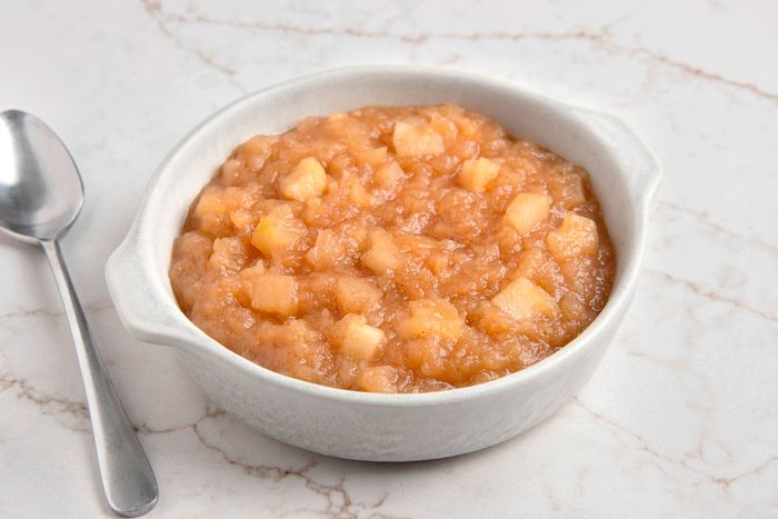 White bowl filled with apple sauce is on a marble surface beside the bowl is a silver spoon