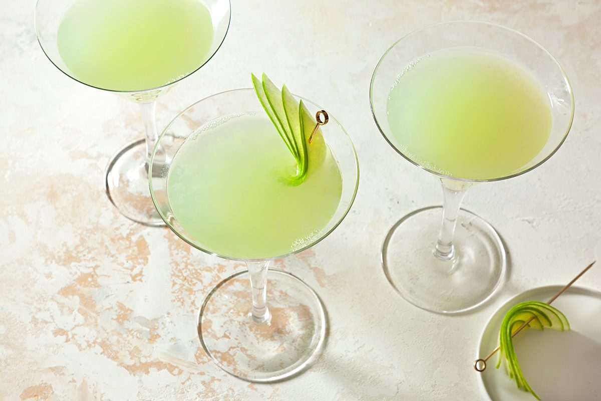 Three martini glasses filled with a light green cocktail are arranged on a textured surface. Each drink has a garnish of thinly sliced green apple skewered on a toothpick.